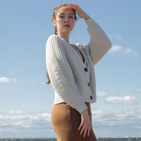 Woman standing on a beach wearing a beige cardigan and brown skirt with a blue sky and ocean background.